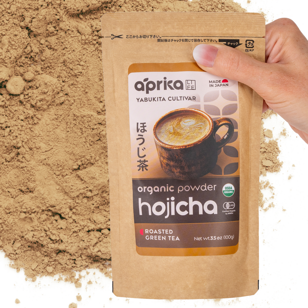 A hand holds a packet of Hojicha Green Tea Powder, made from roasted Japanese tea leaves, with loose powdered tea sprinkled in the background.
