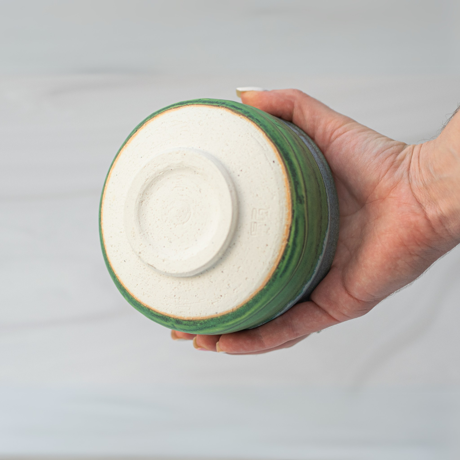 A hand holds the Iguchi Deep Sea Clay Matcha Bowl with a green rim and white base crafted using Minoyaki pottery, against a light background.
