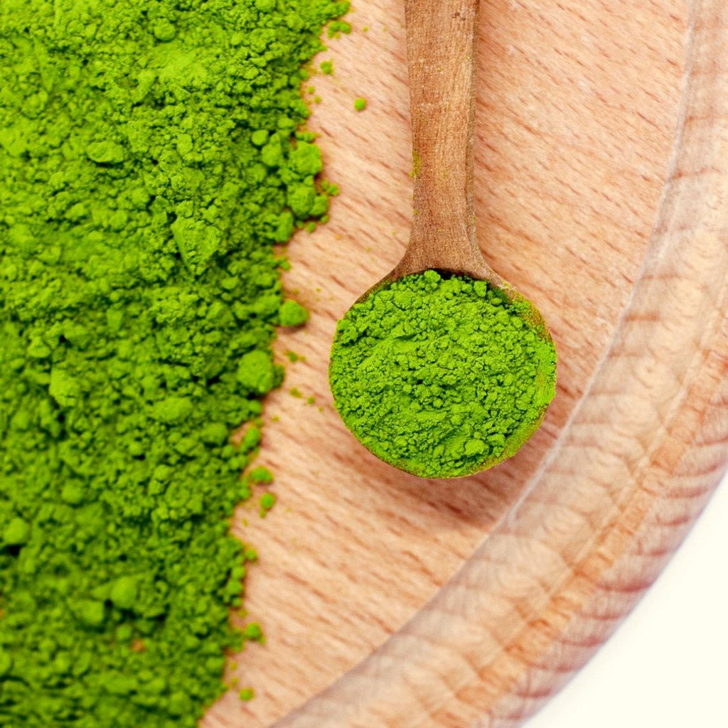 A wooden spoon with Organic Japanese Matcha Green Tea Powder rests on a wooden surface, surrounded by vibrant green matcha powder.