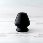A Black Matcha Whisk Holder from the Bamboo Whisk Set, echoing a Japanese aesthetic, sits elegantly on a white wooden surface.