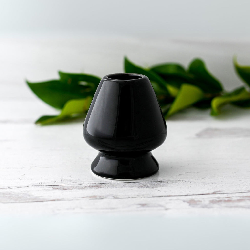 The Black Matcha Whisk Holder from the Bamboo Whisk Set sits on a white surface with green leaves in the background, evoking a serene Japanese aesthetic.