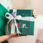 A person holds the Green Unfurl Matcha Gift Set—a green box with "Unfurl," a white ribbon, and flowers—ideal for anyone who enjoys a thoughtful matcha tea ritual.