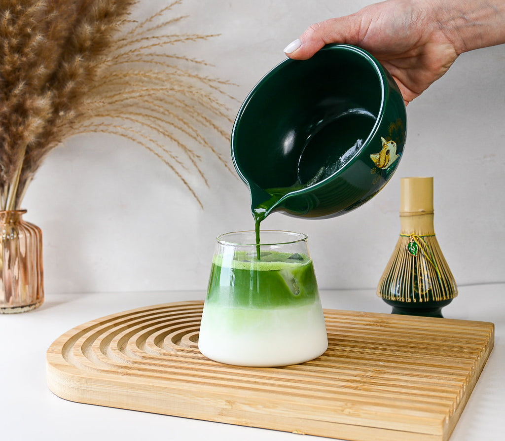 A hand pours Green Unfurl Matcha from the Gift Set bowl into a glass of milk on a wooden tray, with a whisk and vase behind, capturing the essence of the matcha ritual.