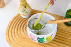 A hand sifts matcha powder into a kawaii cat bowl with cartoon cats using the strainer and bamboo stick from the White Unfurl Matcha Gift Set, all arranged on a wooden tray.
