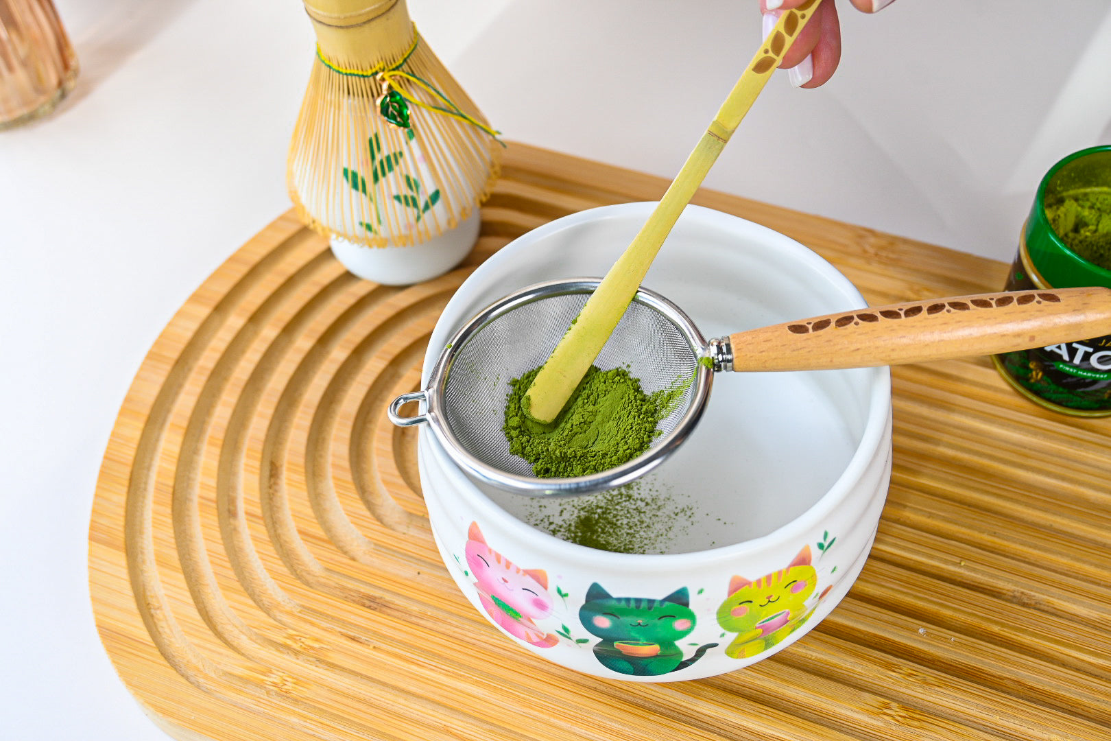 A hand sifts matcha powder into a kawaii cat bowl with cartoon cats using the strainer and bamboo stick from the White Unfurl Matcha Gift Set, all arranged on a wooden tray.