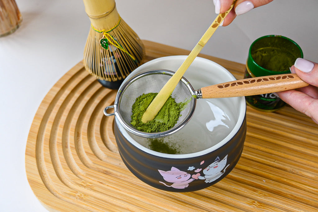 A hand sifts green matcha powder into a kawaii cat bowl on a bamboo tray, ready to be whisked with the Black Unfurl Matcha Gift Set.