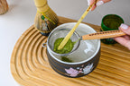 A hand sifts green matcha powder into a kawaii cat bowl on a bamboo tray, ready to be whisked with the Black Unfurl Matcha Gift Set.