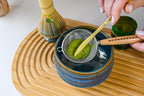 A hand sifts green matcha powder into a blue bowl on a wooden tray, with a whisk nearby—a serene Japanese tea ceremony moment captured with the Ceremonial Matcha Starter Kit - Blue.