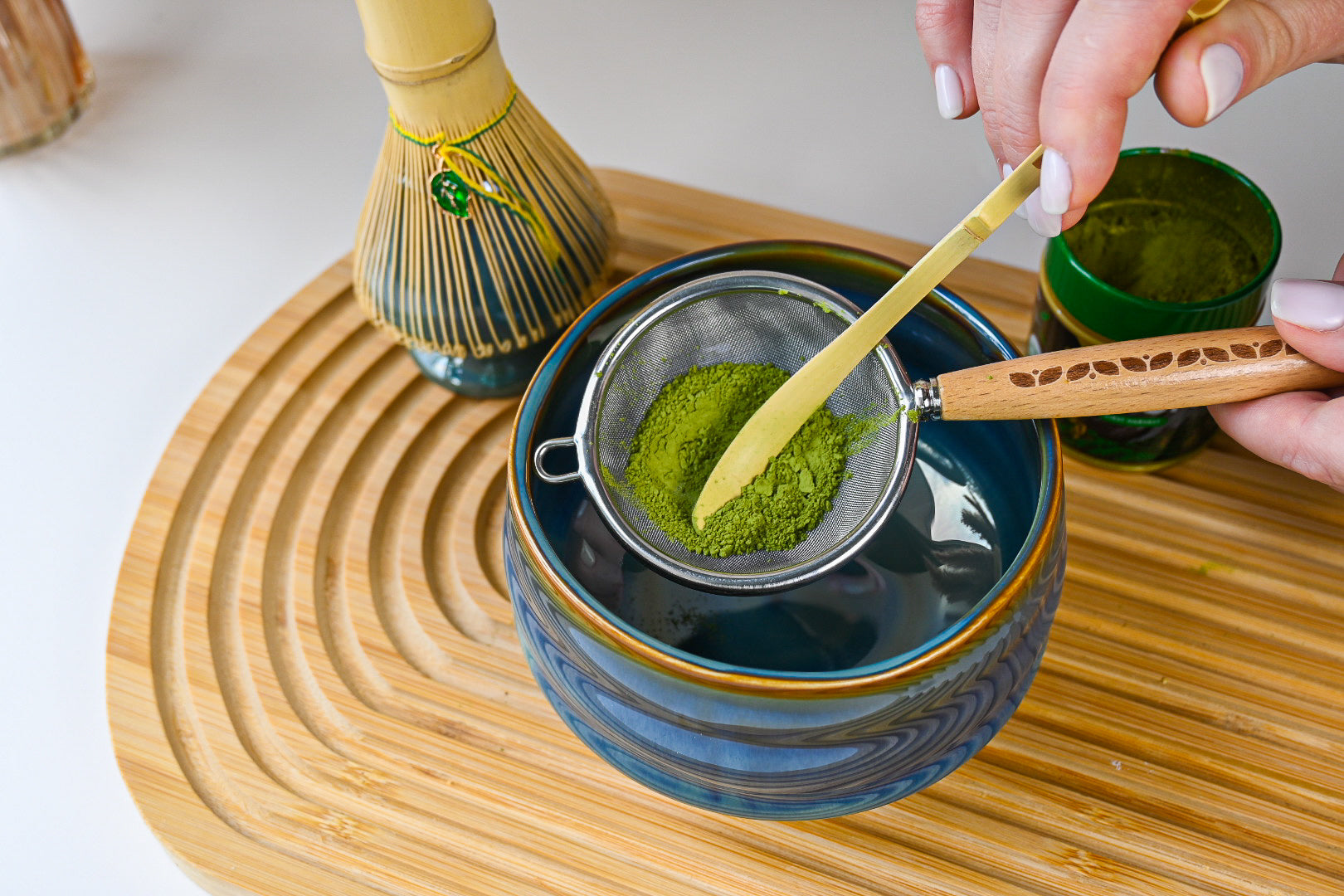 A hand sifts green matcha powder into a blue bowl on a wooden tray, with a whisk nearby—a serene Japanese tea ceremony moment captured with the Ceremonial Matcha Starter Kit - Blue.