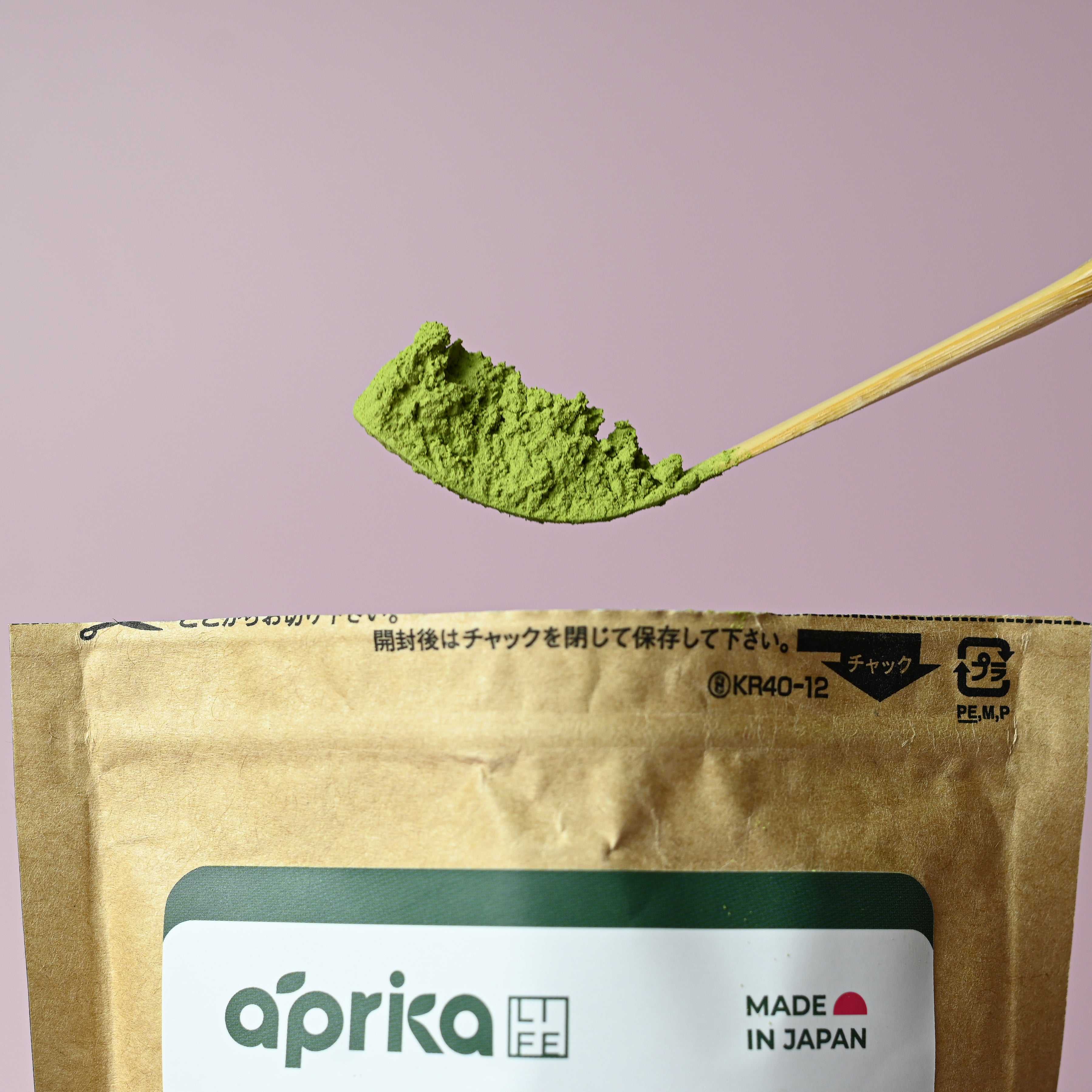 A bamboo spoon with vibrant Culinary Grade Matcha Powder for Latte, Smoothies and Recipes - 3.5 Oz above a brown tea package on a pink background.