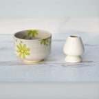 A Green Flowers Ceramic Matcha Bowl sits next to a small white egg cup on a light wooden surface.