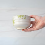 A hand holds the Green Flowers Ceramic Matcha Bowl, a white ceramic bowl adorned with green floral patterns, against a light background.
