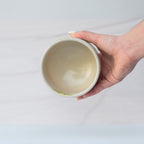 A hand holds the Green Flowers Ceramic Matcha Bowl, crafted from Japanese pottery, above a light, blurred background.