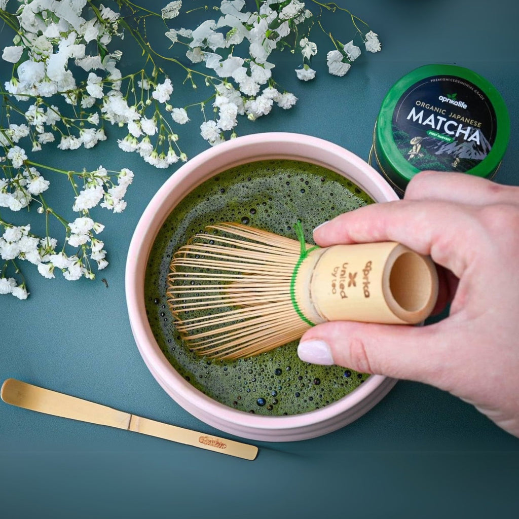 Using the Matcha Bamboo Whisk & Holder Set, enjoy hand whisking matcha in a bowl, surrounded by flowers, with a bamboo scoop nearby and a matcha container elegantly within reach.