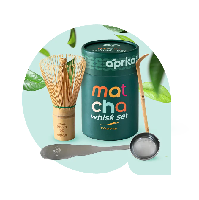 Matcha whisk set with a bamboo whisk, green tin, spoon, and ladle, surrounded by green tea leaves.