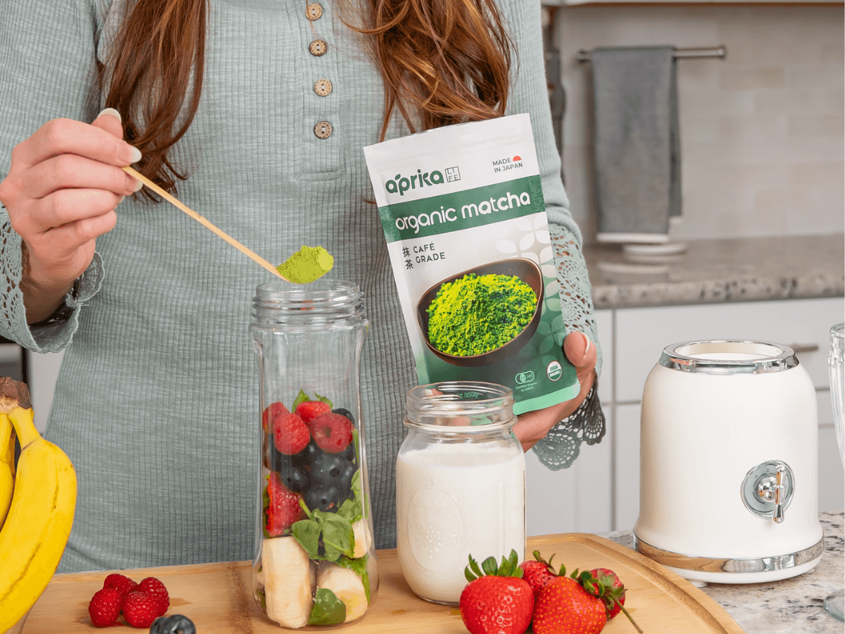 A woman adds matcha powder to a blender with fruit, next to milk and a bag labeled "organic matcha.