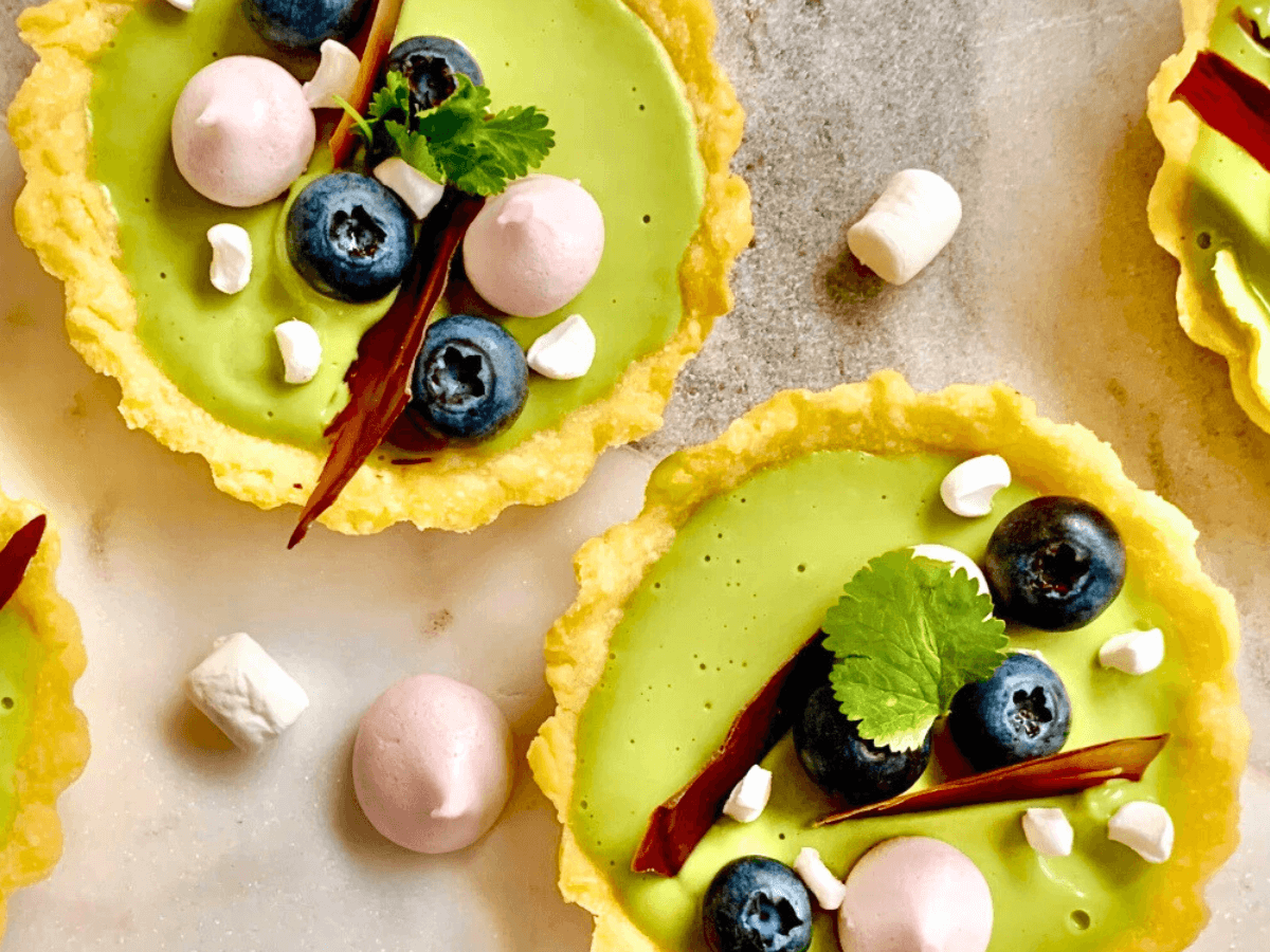 Mini tarts with green filling, blueberries, pink meringue, chocolate sticks, and fresh herbs on a marble surface.