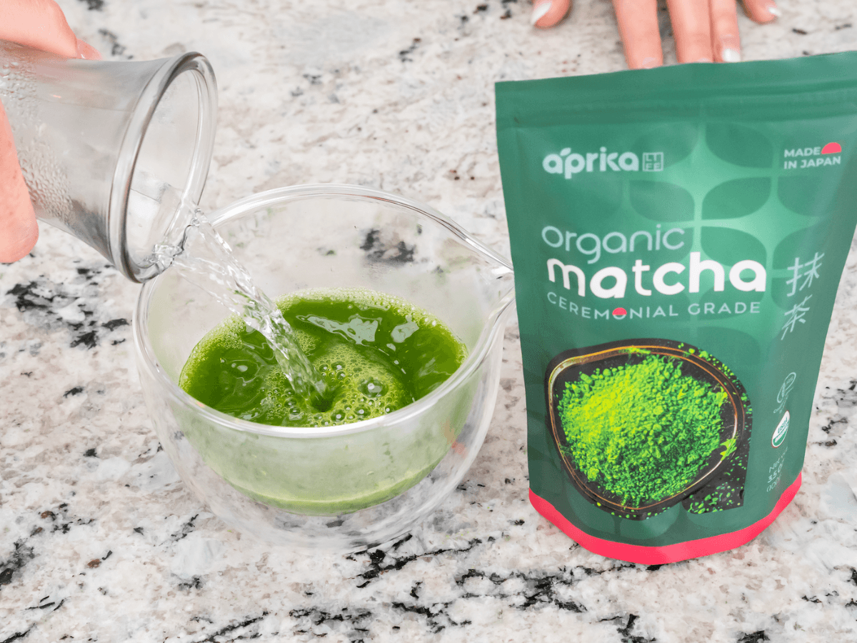 A hand pours water into a bowl of matcha powder beside a bag labeled "organic matcha ceremonial grade.