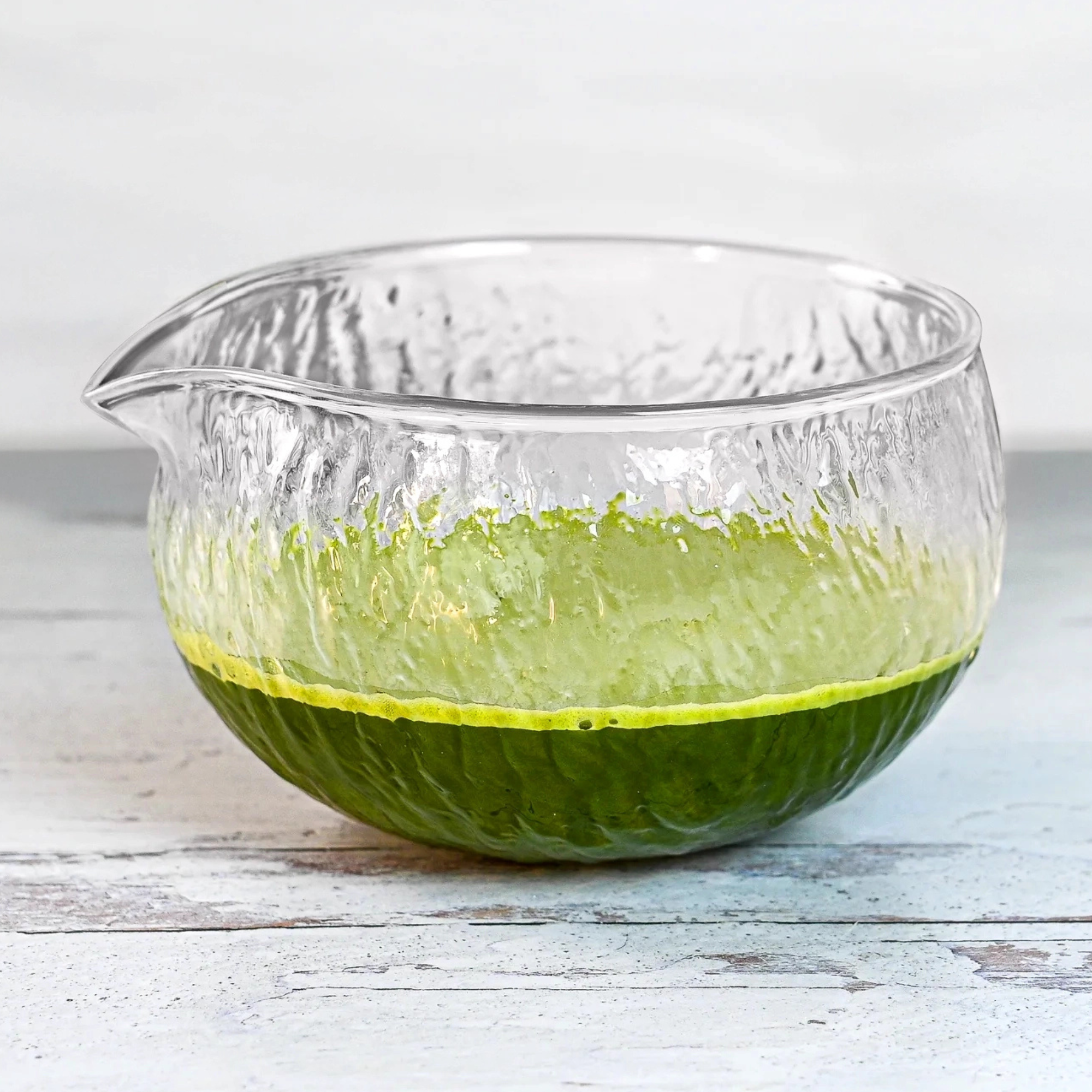 The Textured Spouted Glass Matcha Bowl Pattern 2, holding vibrant green matcha, sits elegantly on a white wooden surface.