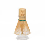 The Clear Glass Matcha Whisk Holder and Bamboo Whisk Set is shown with the bamboo whisk resting on the glass holder against a white background, making it an ideal addition to any matcha tea set.