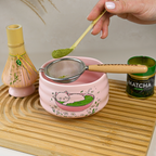 A hand sifts matcha powder into a Cat Print Bowl, part of the Pink Unfurl Matcha Gift Set, with a whisk and matcha tin on a wooden tray.