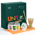The White Unfurl Matcha Gift Set includes a kawaii cat bowl, cute cat cup, strainer, whisk, scoop, bowl, and orange cloth beside a green gift box—ideal as a white matcha gift set.